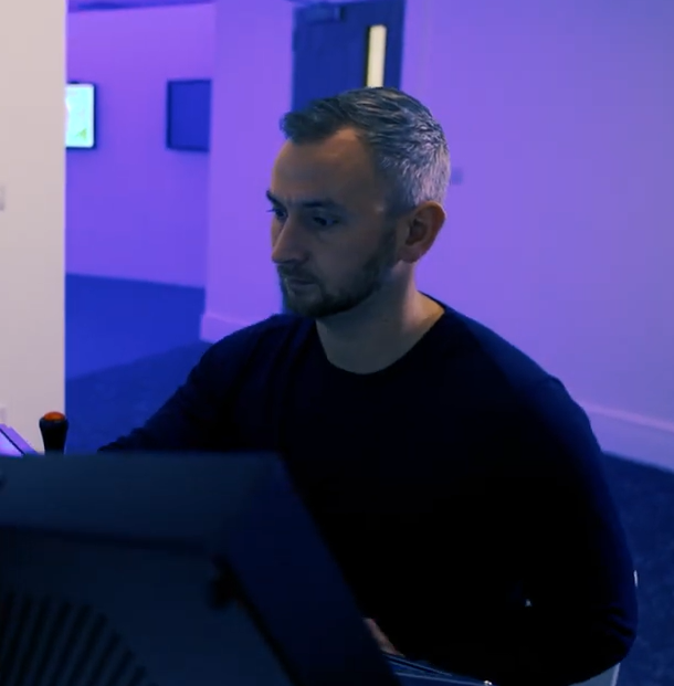 A man with short hair and a beard sits at a computer in a room with blue lighting.