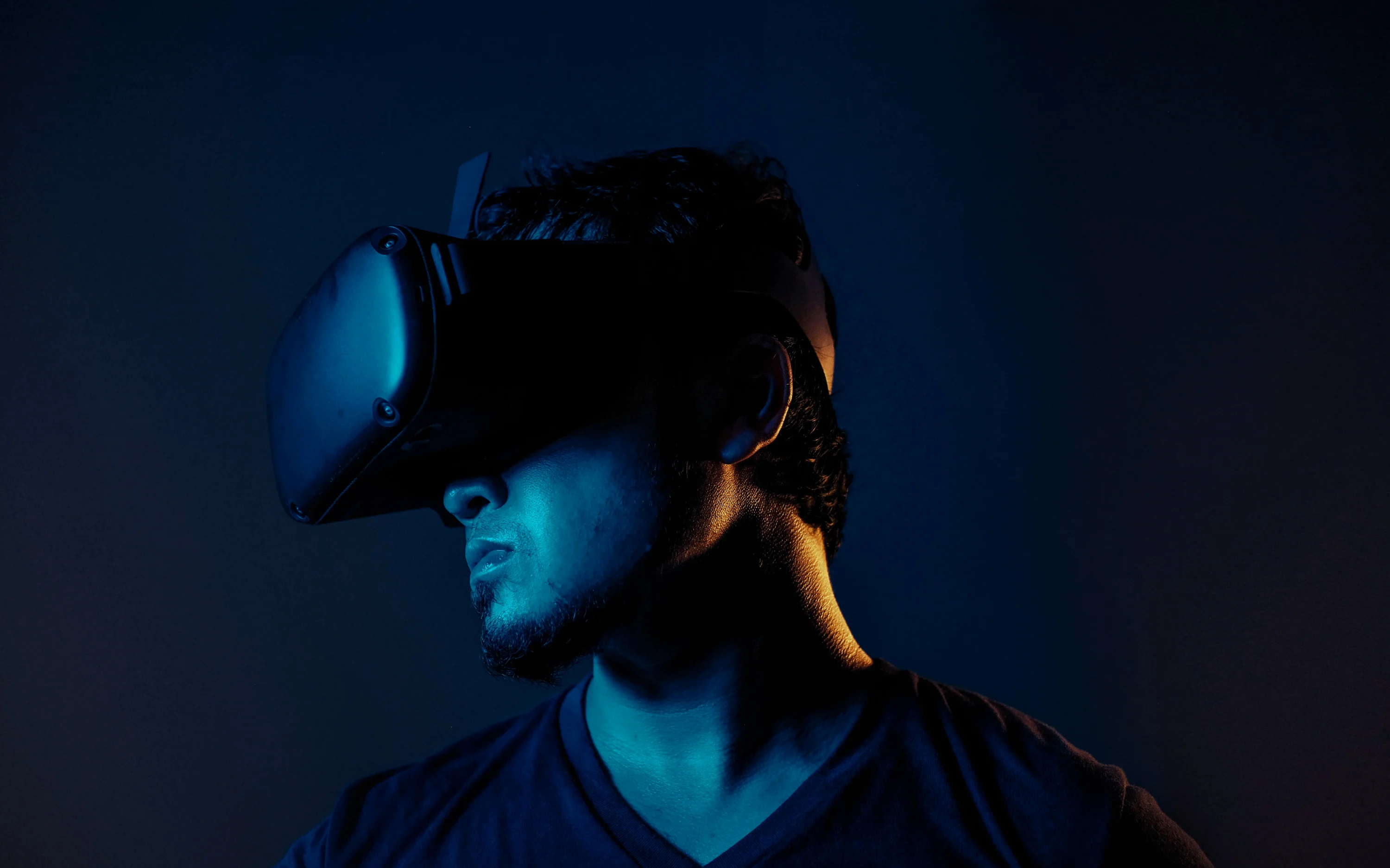 A man with short hair and a beard wearing a virtual reality headset, illuminated with blue light against a dark background.