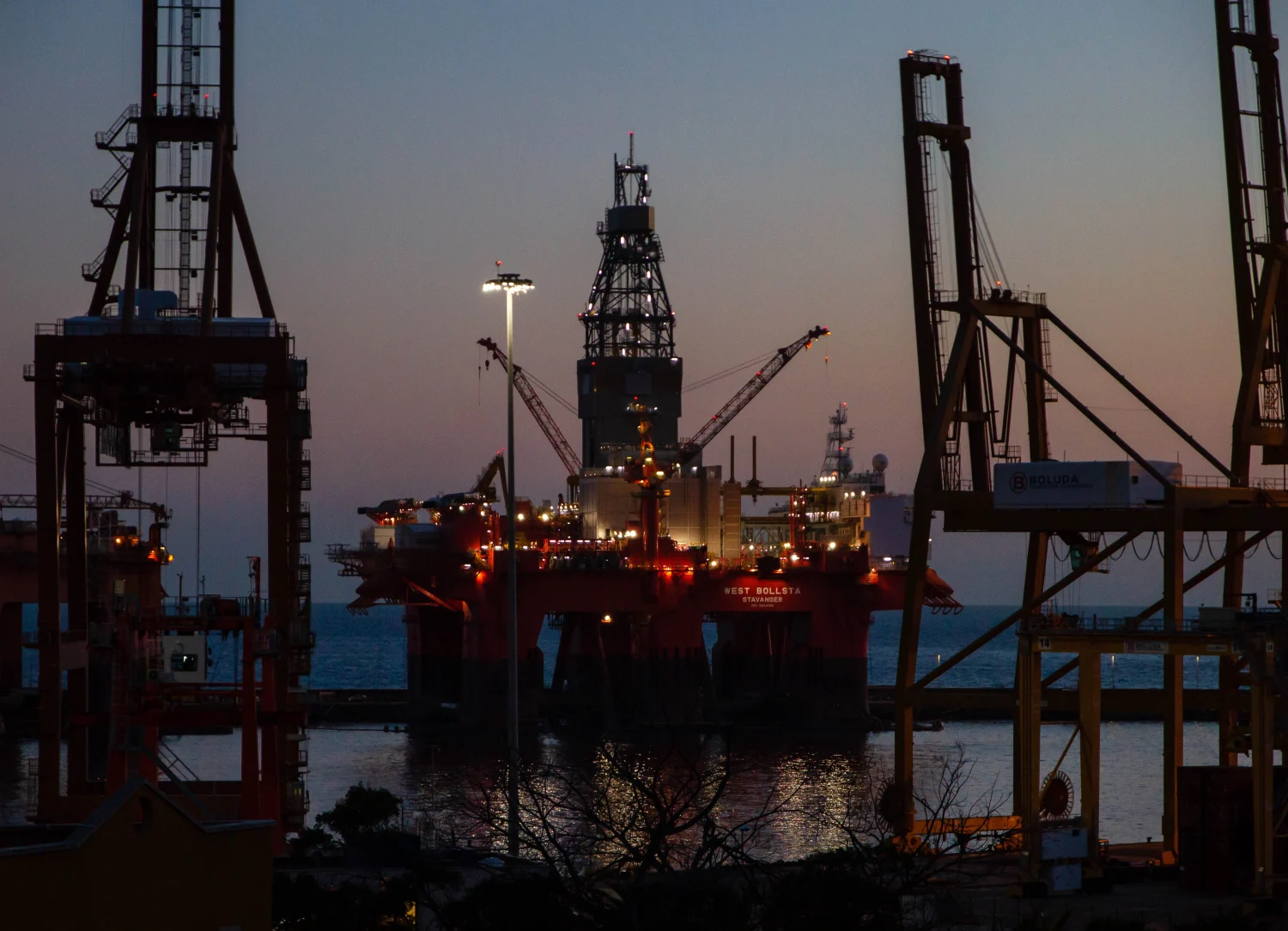 An offshore oil rig lit up at dusk, surrounded by the silhouettes of large cranes and industrial structures, with the sea in the background.