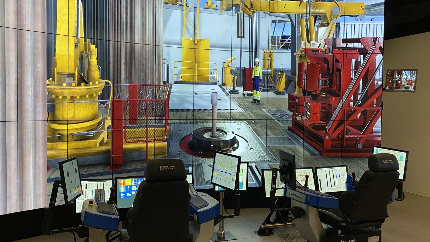 Control room setup with multiple screens and workstations displaying a detailed drilling rig simulation. Operators' chairs and monitors are in the foreground, while the virtual rig scene is in the background.