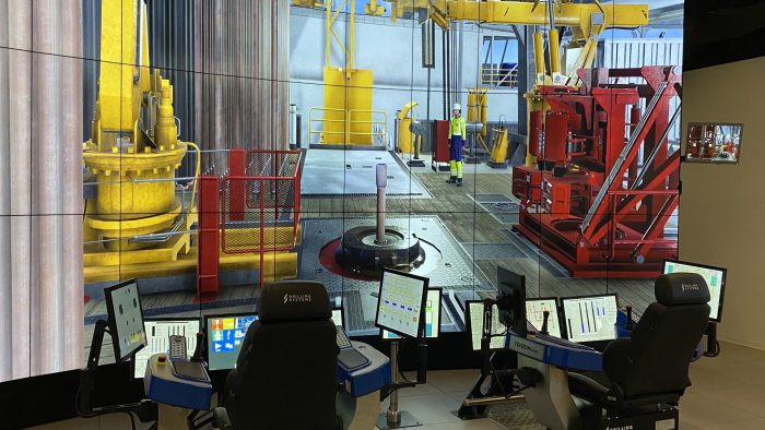 Control room setup with multiple screens and workstations displaying a detailed drilling rig simulation. Operators' chairs and monitors are in the foreground, while the virtual rig scene is in the background.