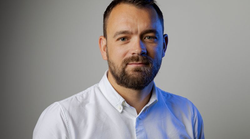 A man with a short beard and short hair wearing a white shirt stands against a plain gray background, looking at the camera.