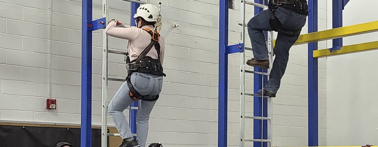 Two individuals in safety gear and helmets are climbing vertical ladders inside an industrial training facility.