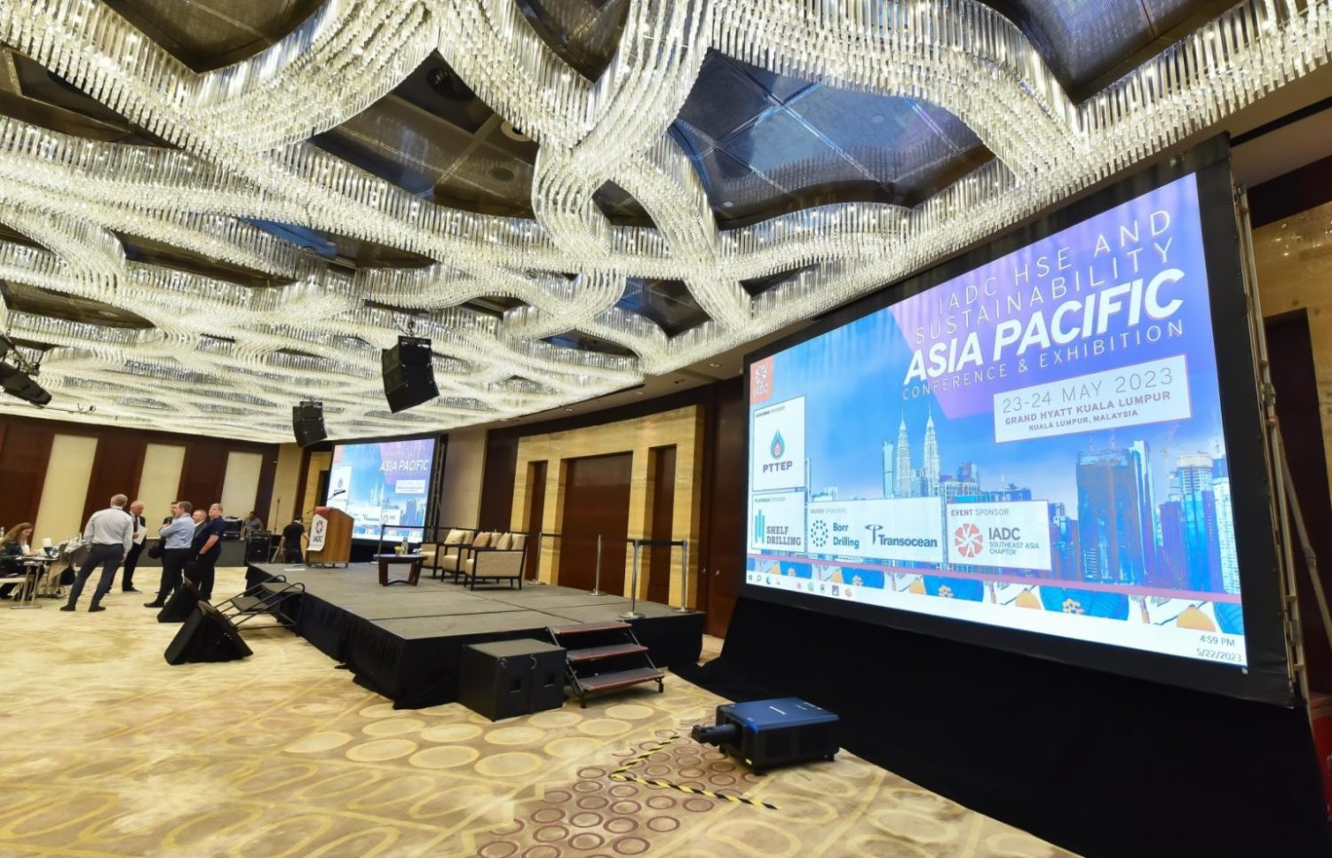 A conference room with attendees facing a stage and large screen displaying details of the 