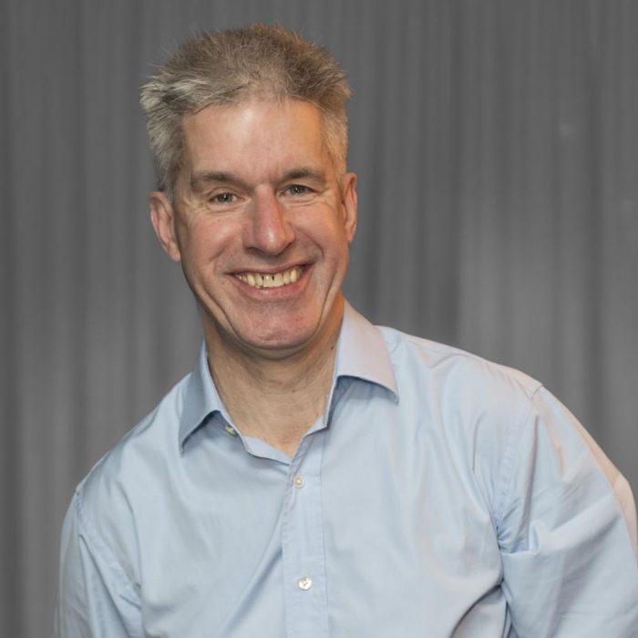 A man with gray hair and a light blue shirt smiles at the camera against a neutral background.