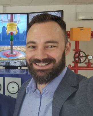 A man with a beard and short hair is smiling in front of a background with a television screen showing a brightly colored industrial scene and red industrial equipment.