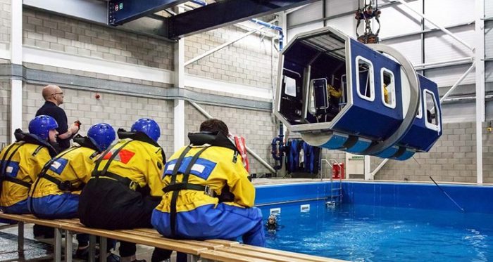 A group of people in yellow and blue safety gear sit on benches, observing a simulated helicopter cockpit suspended above a pool in an indoor training facility.