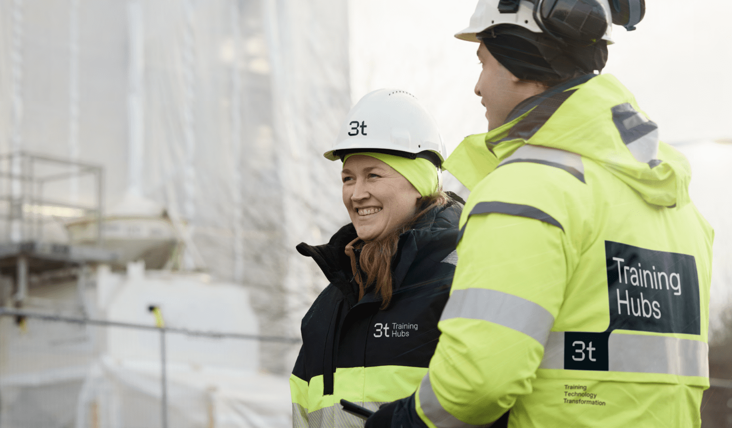 Two workers in high-visibility jackets and helmets stand outdoors near a construction site. Both jackets display the 