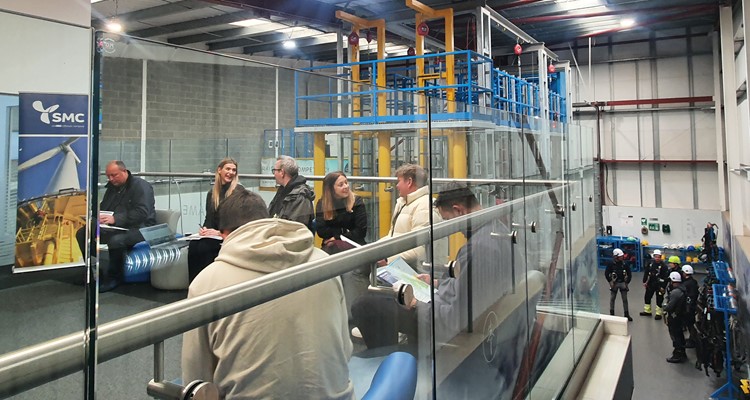 A group of people sits and converses in a modern industrial facility with glass partitions. Safety equipment and machinery are visible in the background.