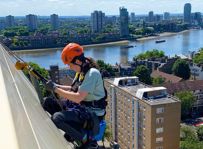 Person wearing safety gear is cleaning the windows of a tall building with a river and cityscape in the background.
