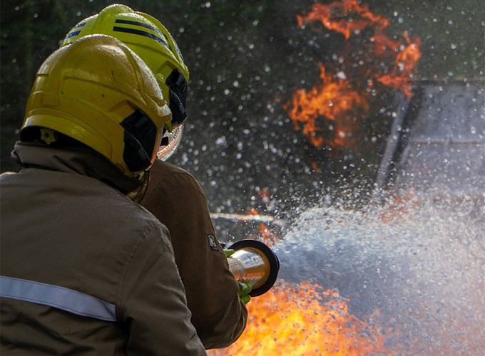 Two firefighters in yellow helmets spraying water at an intense fire with visible flames.