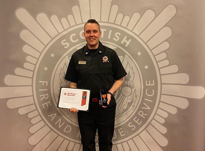 A person in a dark uniform holding a certificate and a medal, standing in front of a Scottish Fire and Rescue Service backdrop.