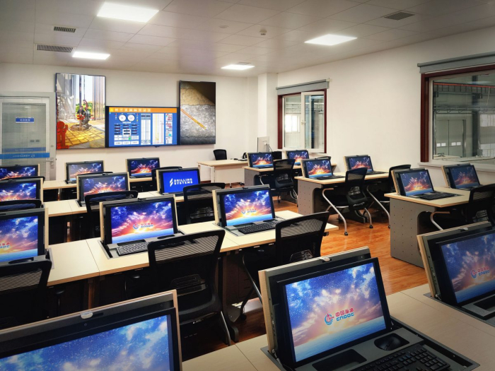 A modern classroom equipped with numerous workstations, each featuring a computer displaying a colorful screensaver. A large digital screen and whiteboard are visible at the front of the room.