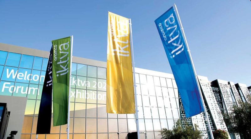 Three vertical flags in green, yellow, and blue stand outside a modern building with glass windows and a welcome sign for the 