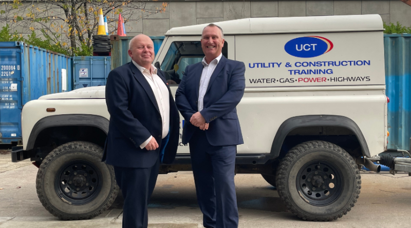 Two men in suits stand in front of a Utility & Construction Training vehicle, which has branding on the side indicating services related to water, gas, power, and highways.