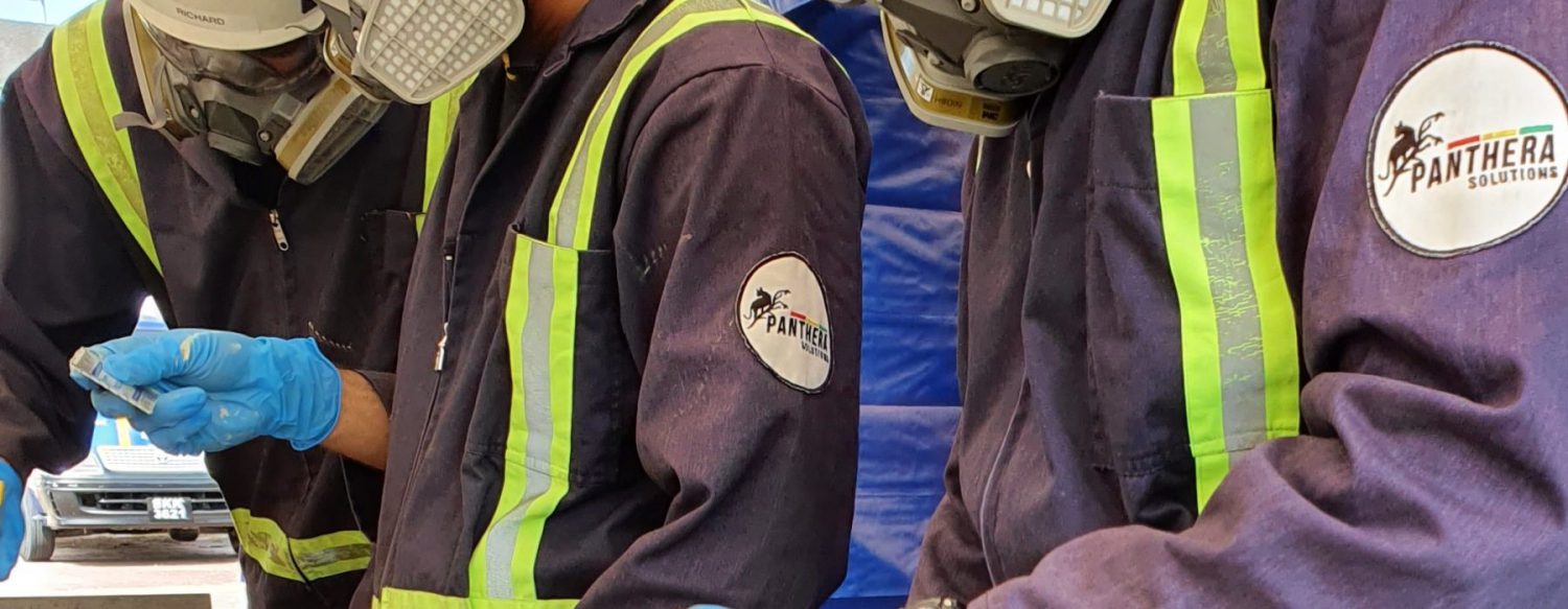 Three individuals wearing protective gear, including face masks and gloves, working closely together. Their dark blue uniforms feature bright green safety stripes and a patch reading 