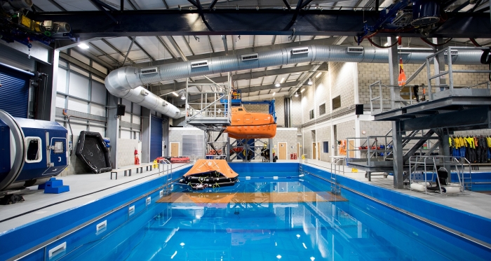 A large indoor facility featuring a pool, with a suspended lifeboat and life rafts on the water. The area is equipped with various apparatus, industrial piping, and a mock airplane door on the left.