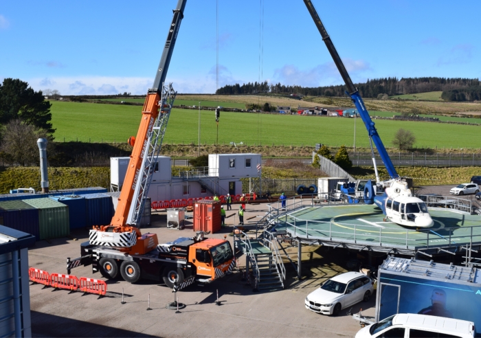 Two cranes operate at a construction site with a helicopter on a platform. Several vehicles and shipping containers are around, with personnel in safety gear working in the area under clear skies.