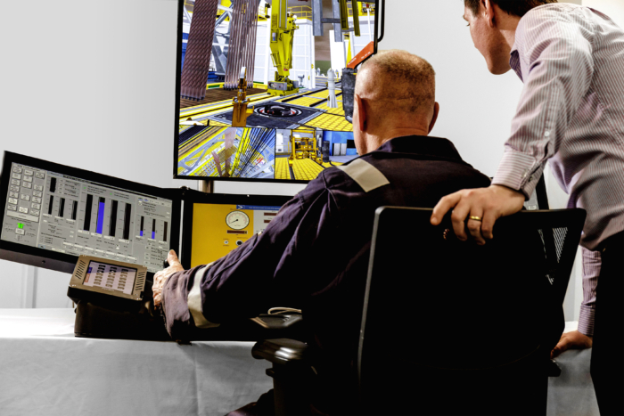 Two individuals observe and interact with data and simulations on multiple monitors in a control room setting. The scene includes industrial equipment displayed on the screens.