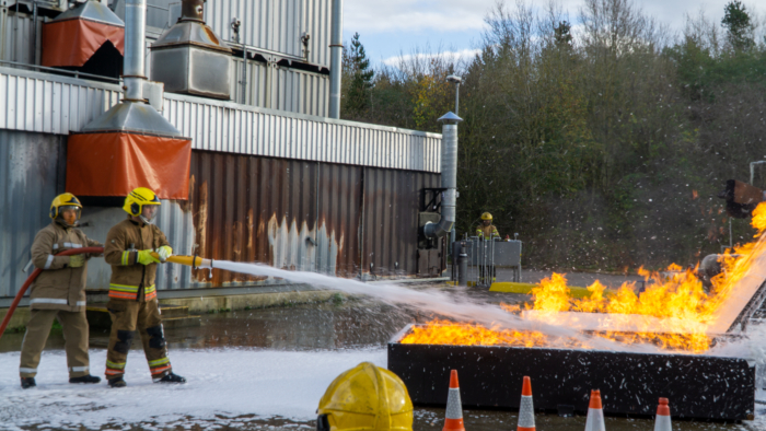Firefighters in protective gear use a large hose to spray foam on a controlled fire outdoors near an industrial facility surrounded by trees. Orange traffic cones are placed in the foreground.