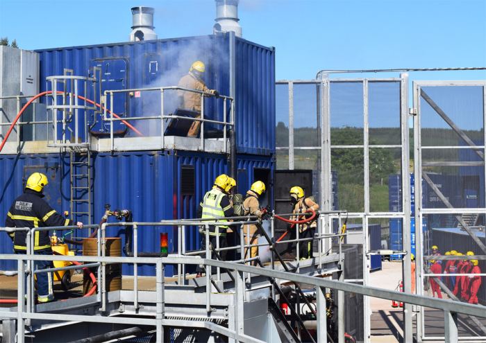 Firefighters in yellow and black uniforms conduct a training exercise, using hoses and equipment on a multi-level blue structure. Smoke is visible from the upper levels.
