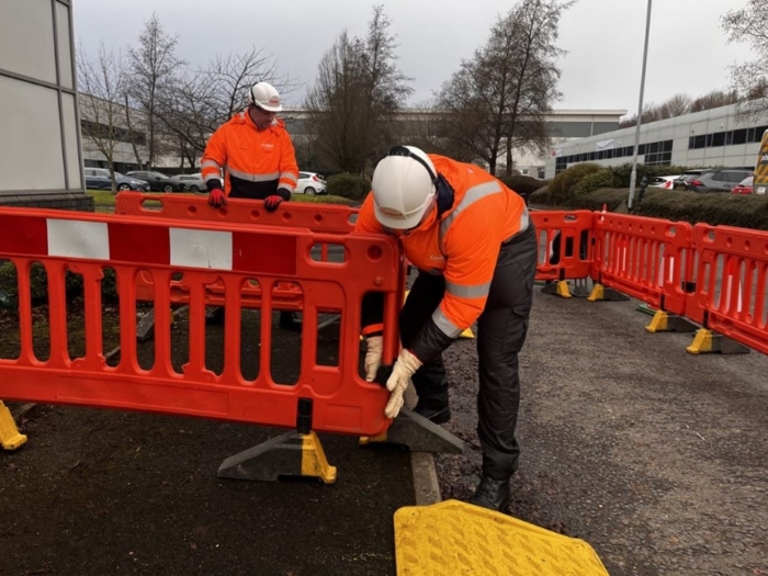 Two construction workers in high-visibility clothing and hard hats are setting up red plastic barriers on an outdoor worksite.