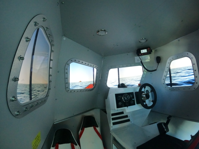 Interior view of a small, metal boat cabin with windows showing an ocean scene. The cabin contains navigation controls, a steering wheel, and seats.
