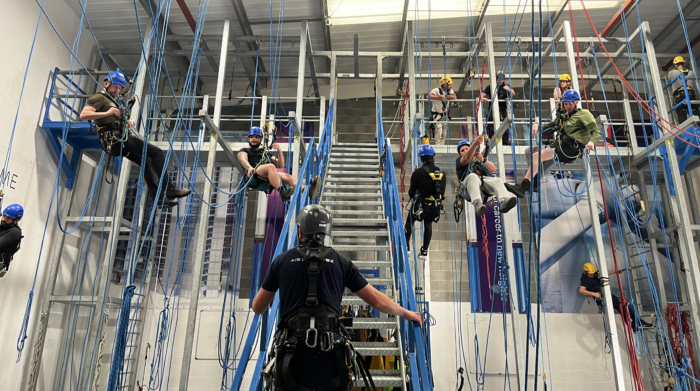 A group of individuals wearing helmets and harnesses participate in an indoor climbing and rope safety training session, supervised by an instructor.