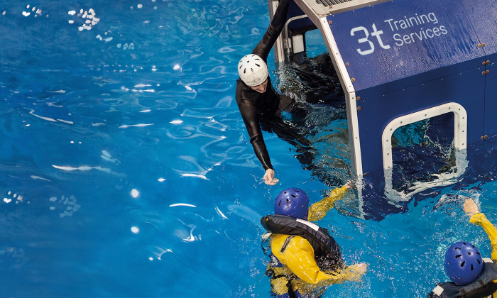 Two individuals in yellow waterproof outfits and helmets receive assistance from a person in a wetsuit and white helmet, emerging from a blue training module labeled 