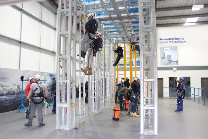 People wearing safety gear are practicing climbing and rescue techniques on metal structures inside an indoor training facility labeled 