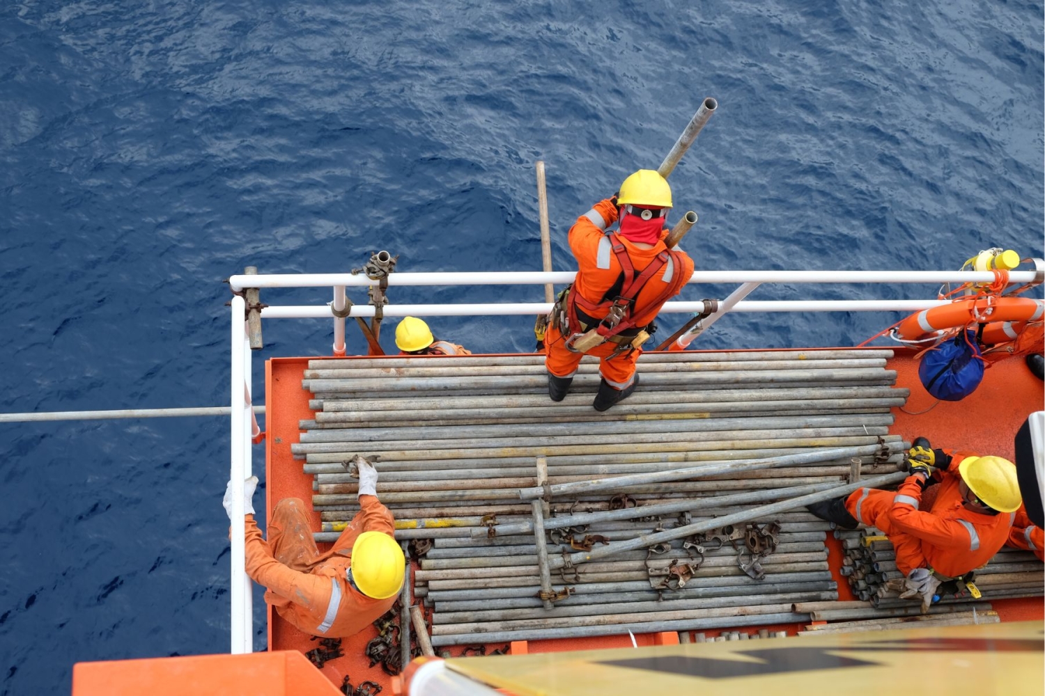 Workers in orange safety gear and yellow helmets handle metal pipes on the deck of an offshore platform against a backdrop of the ocean.