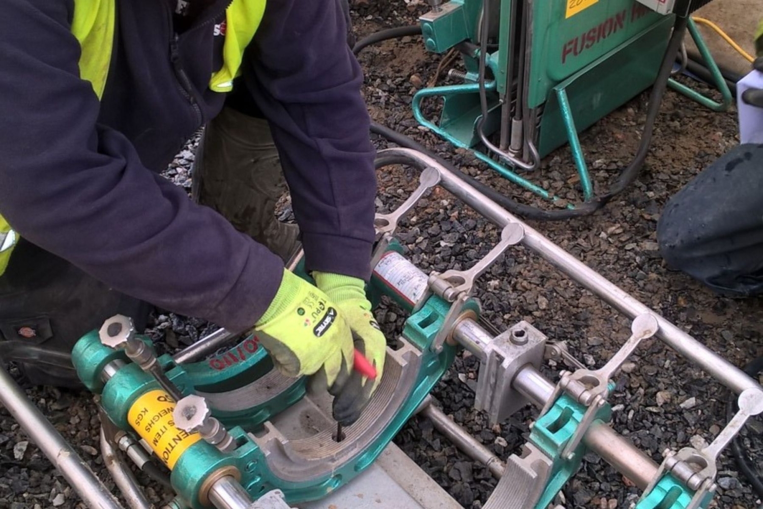 A person wearing safety gloves and a high-visibility jacket is using a wrench on a large, green pipe fusion machine outdoors on a gravel surface.