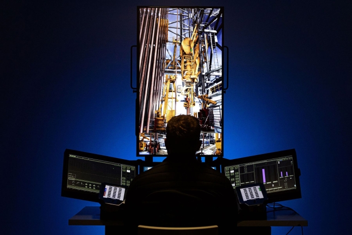 A person sits at a control station with multiple monitors displaying industrial machinery, suggesting an operation or monitoring of equipment.