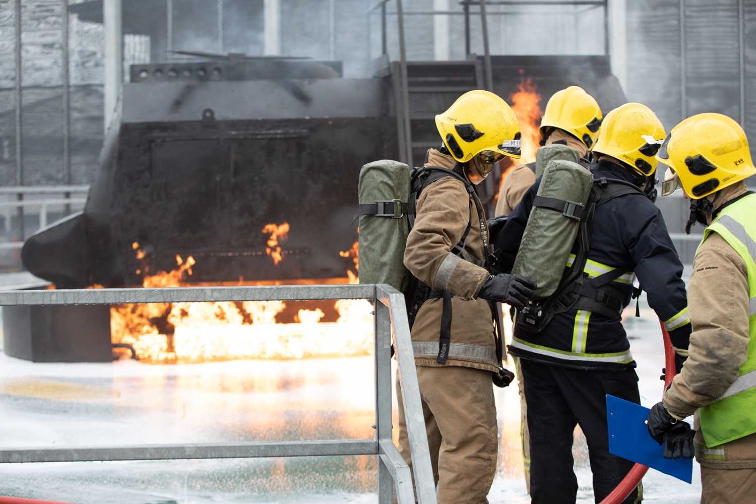 Firefighters in yellow helmets work to extinguish a fire near a damaged, burning structure. They are using hoses and carrying equipment, while flames and smoke are visible in the background.