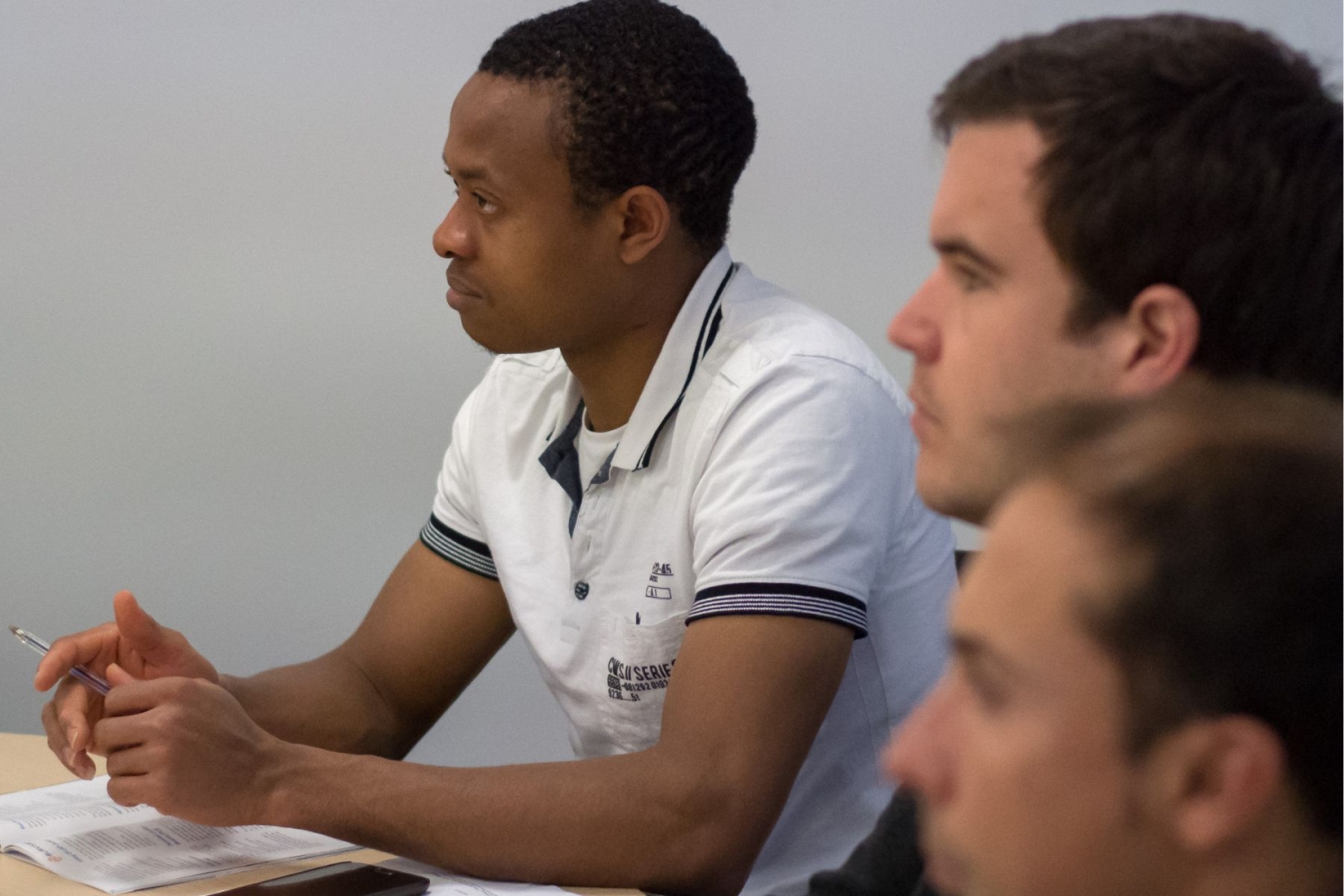 Three men sit at a table, attentively focusing on something off-camera. The man on the left holds a pen and wears a white collared shirt, and they are engaged in a discussion or meeting.