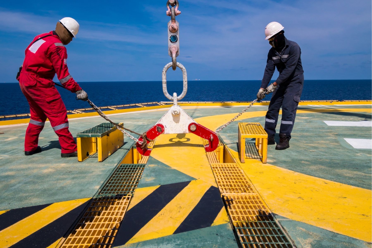 Two workers in safety gear adjust heavy-duty chains on a helideck against a backdrop of the ocean and blue sky.