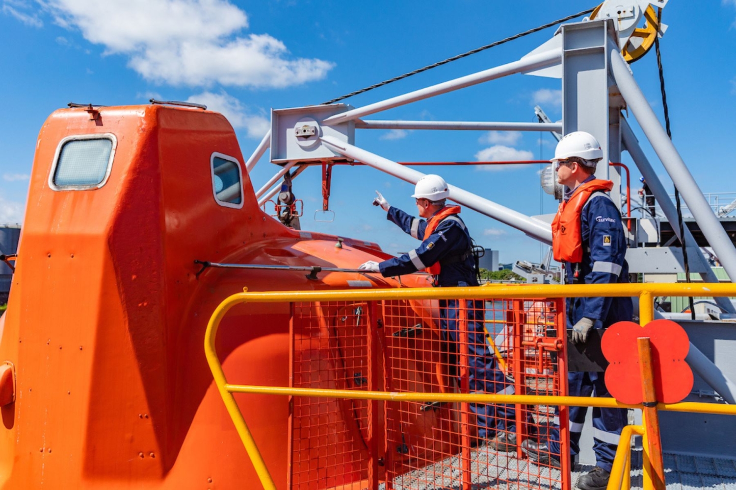 Two workers in safety gear inspect an orange lifeboat on the deck of a ship under a clear blue sky.