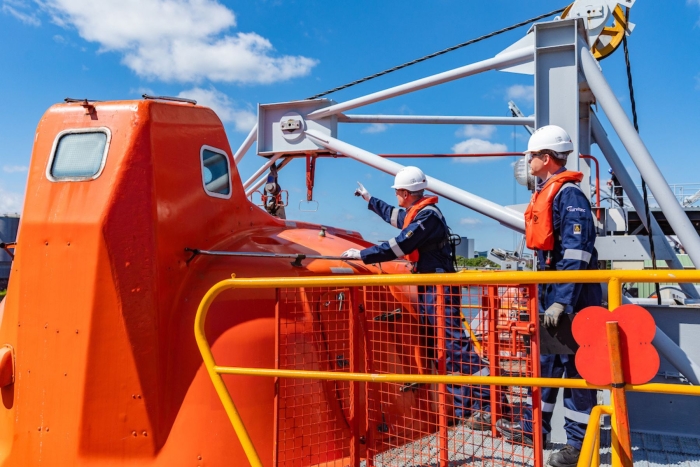 Two workers in safety gear inspect an orange lifeboat on the deck of a ship under a clear blue sky.