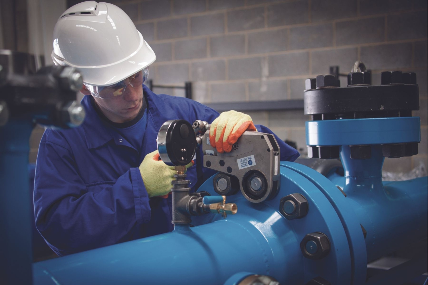 A technician wearing safety gear adjusts a piece of equipment on a large blue industrial pipe in a facility.
