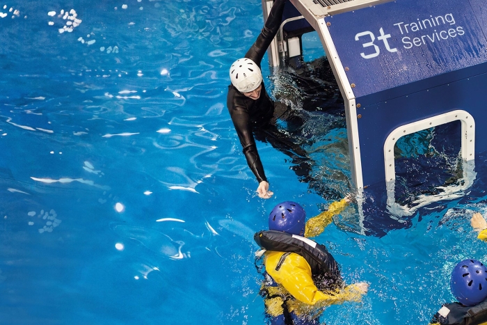 Two individuals in the water perform a safety exercise with one person reaching out from a cabin labeled 