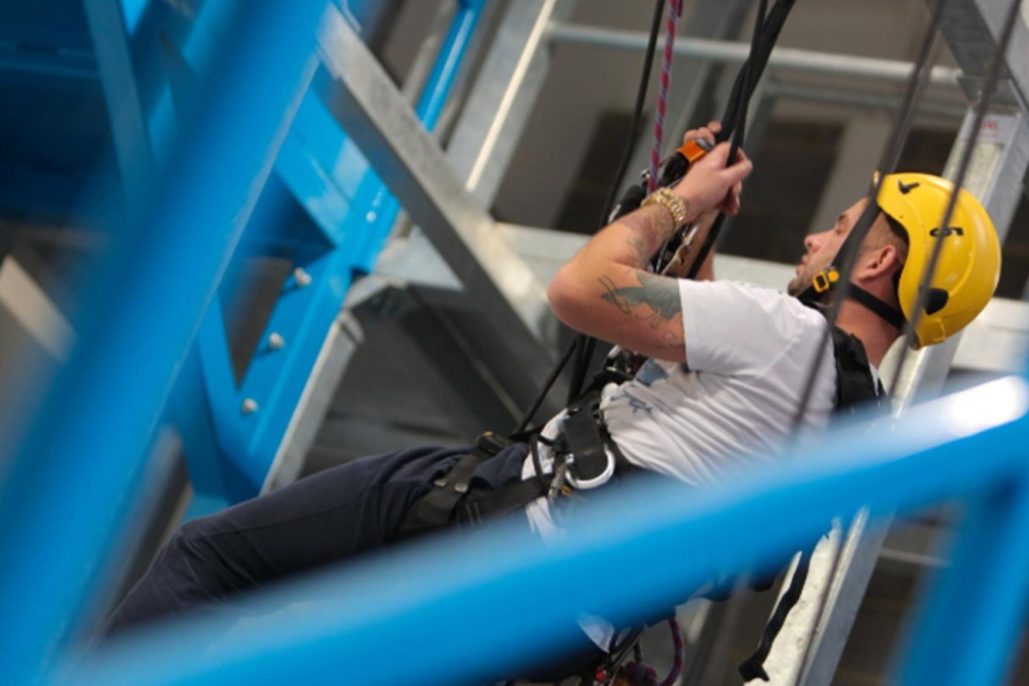 Person wearing a yellow helmet and harness performing an indoor climbing or industrial safety training exercise with blue metal structures in the background.