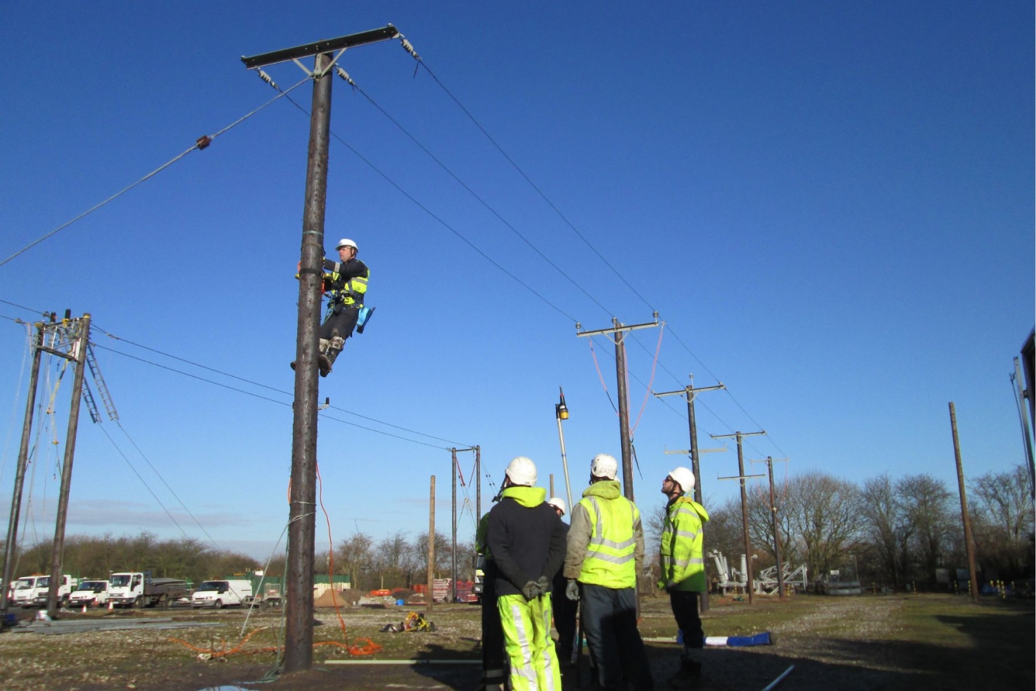 Worker wearing safety gear climbs a utility pole while four others in high-visibility clothing and helmets observe from the ground on a clear day.