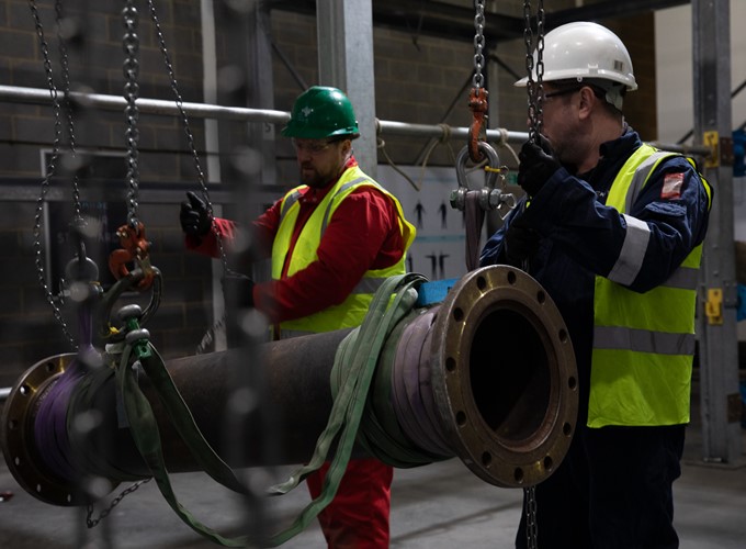 Two workers in safety vests and helmets lift a large pipe using a chain hoist in an industrial setting.