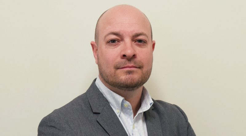 A bald man with a beard is wearing a grey suit and light-colored shirt, standing against a plain background.
