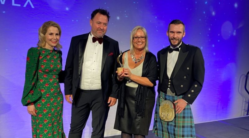 Four people at an event, with one woman in the center holding a trophy. Three individuals are wearing formal attire, including tuxedos and a kilt, while the woman on the left wears a green dress with red accents.