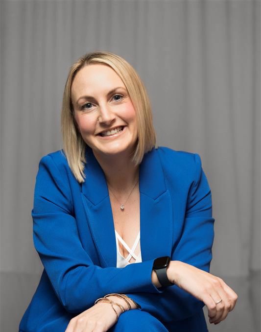 A person wearing a blue suit and a smartwatch, smiling while seated with arms resting on knees, and a gray curtain in the background.