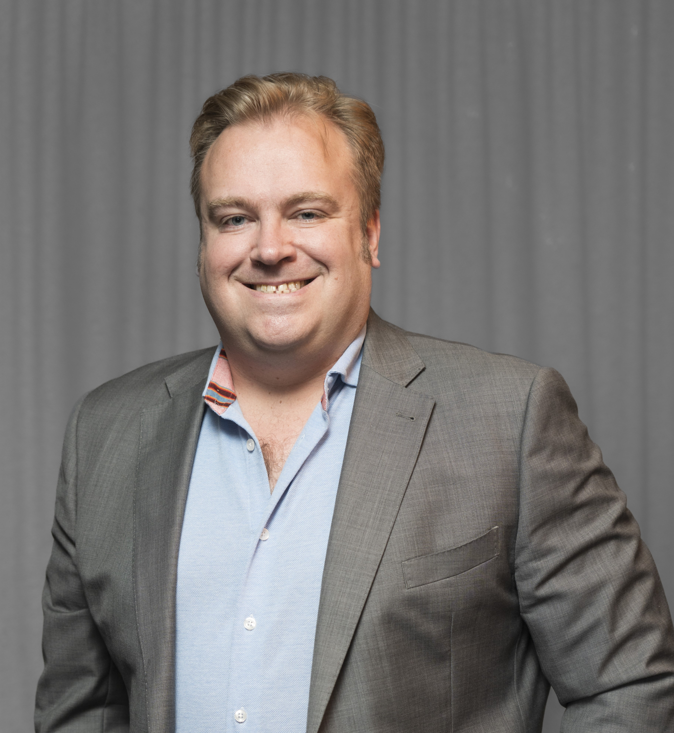 A man with blond hair is smiling, wearing a grey suit jacket over a light blue shirt, standing in front of a grey curtain.