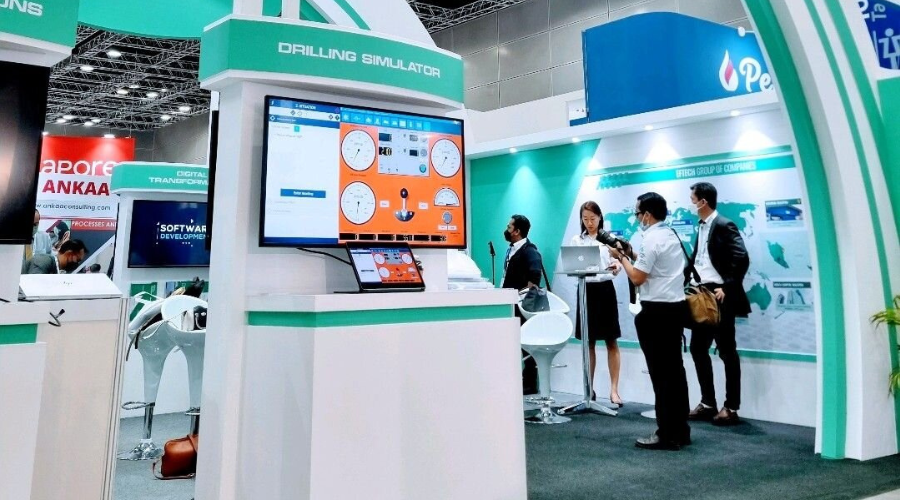 People interacting at a trade show booth featuring a drilling simulator display, with computer screens and informational panels.