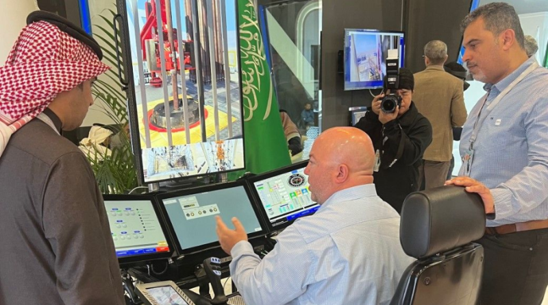 Three men are engaged in conversation near a control panel with multiple screens. One man, wearing a headscarf, faces a seated man who gestures toward the screens. Another man stands nearby.