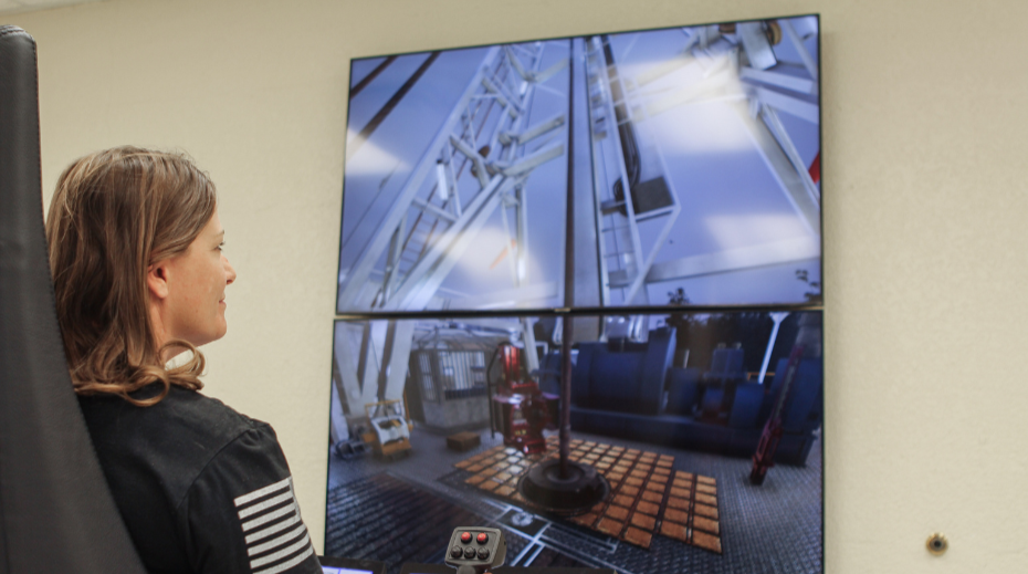 A woman operates a control panel while watching a large screen displaying machinery in an industrial setting.